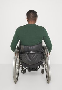 Grön långärmad skjorta bärs av en person i rullstol, som har en mörk dämpad ryggstöd, silverfärgade ekrar och svarta hjul.