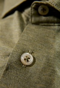 Das khakifarbene Hemd mit durchgehender Knopfleiste besteht aus einem glatten, strukturierten Stoff und ist mit einem perlmuttartigen Knopf versehen, der ein Vierlochdesign und kontrastierende Nähte zeigt.