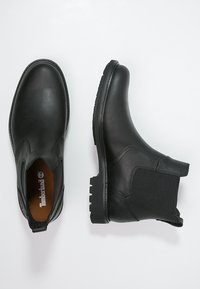 Botas de tobillo de cuero negro con paneles laterales elásticos y suelas de goma texturizadas. Presenta un diseño minimalista sin logotipos visibles en el exterior.