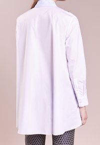 Biała koszula z długim rękawem i złożonym niebieskim, paskowanym kołnierzykiem. Tył ma prosty krój, wykonany z gładkiego, lekkiego materiału.