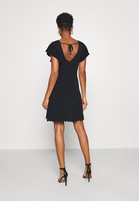 Robe noire avec un décolleté arrondi au dos et de courtes manches volantées. Ourlet à volants. Portée avec des sandales à talons hauts noires à lanières.