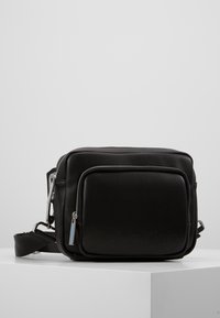 Czarna skórzana torba crossbody z przednią kieszenią na zamek błyskawiczny, srebrnymi uchwytami zamka i regulowanym paskiem na ramię, położona na białej powierzchni.