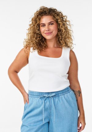 Krullendharige vrouw in een wit mouwloos topje en lichtblauwe broek met koordsluiting, glimlachend met hand in de zak, bloemen-tatoeage op de onderarm.