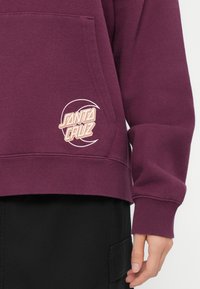 Felpa bordeaux con tasca a marsupio, polsini e orlo a coste. Presenta un logo "SANTA CRUZ" ricamato in beige sul lato.