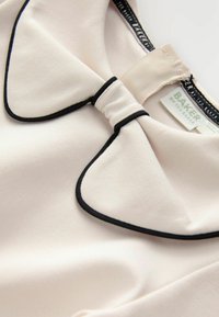 Tessuto rosa chiaro con una texture liscia, caratterizzato da un colletto con rifinitura nera e un dettaglio a fiocco; etichetta visibile che recita "BAKER di Ted Baker."