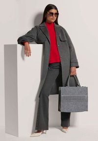 Grau Wolle Mantel über einem roten Pullover, kombiniert mit dunklen Jeans und cremefarbenen Pumps. Eine graue Filztasche mit "MADELEINE" eingeprägt auf der Vorderseite.