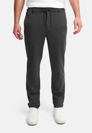 Man draagt donkergrijze broek met trekkoord en witte sneakers, staand tegen een lichte achtergrond.