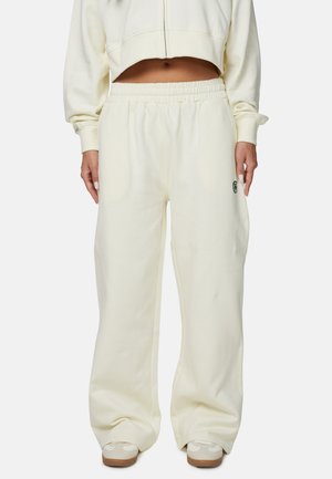 Kremowe, luźne spodnie dresowe z elastycznym paskiem w talii, bocznymi kieszeniami i subtelnym logo po lewej stronie. Miękka tekstura materiału.