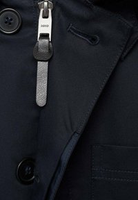 Tessuto trench coat blu navy con grande bottone, zip nascosta e dettaglio in pelle. Presenta linee pulite e una finitura texture.