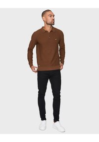 Brauner gestrickter Polohemd mit langen Ärmeln, Kragen und gerippten Bündchen. Hat ein kleines grünes Emblem und wird kombiniert mit schwarzen Jeans und weißen Sneakern.