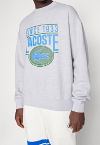 Personne portant un sweat-shirt Lacoste gris clair avec un logo bleu et vert, et un pantalon blanc avec des rayures bleues et un petit emblème.