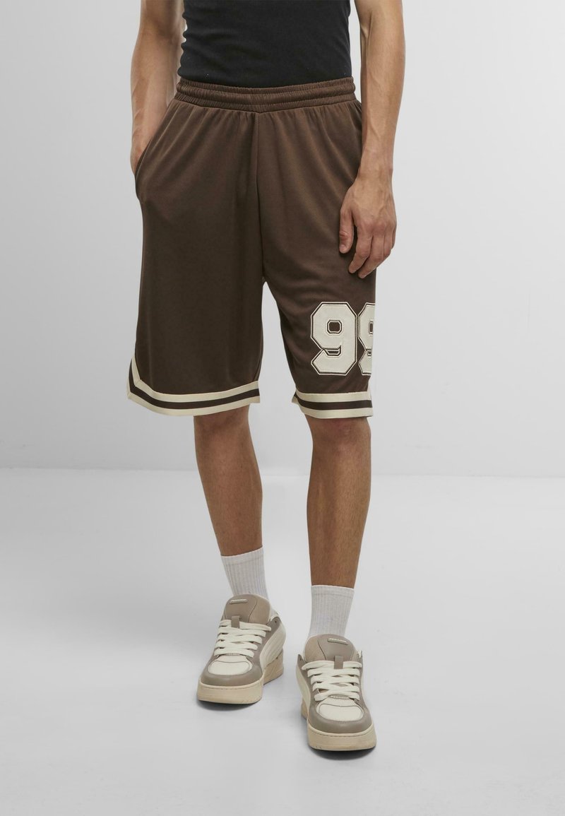 Shorts de sport marron avec des accents blancs, portant le numéro 99, une taille élastique et des poches latérales. Portés avec des baskets beiges et des chaussettes blanches.
