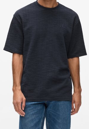 Mand iført en mørk tekstureret kortærmet skjorte med lille logo på brystet og blå denimjeans, stående mod en ensfarvet baggrund.