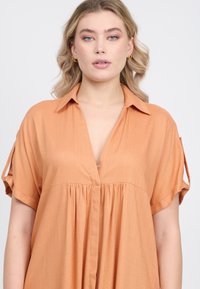 Camisa de color durazno, de manga corta, con cuello y escote en V, que presenta un diseño de frente plisado y tejido texturizado.