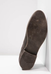 Suela de zapato negro de vestir con patrón de desgaste, talla 42, posicionada verticalmente contra una superficie blanca en un rincón.