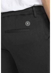 Les pantalons en tissu noir présentent une poche arrière avec un rabat rectangulaire et un accent de logo circulaire. Le matériau a une texture lisse.