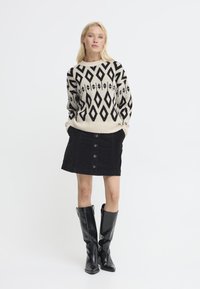 Donna che indossa una gonna nera con bottoni, stivali neri fino al ginocchio e un maglione crema con un motivo geometrico a diamante nero, che sta in piedi con le mani nelle tasche.