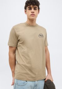 Νεαρός άνδρας που φοράει μπεζ γραφικό μπλουζάκι και ανοιχτό μπλε τζιν, κρατώντας ένα σκούρο μπουφάν μπροστά σε έναν απλό ανοιχτό φόντο.