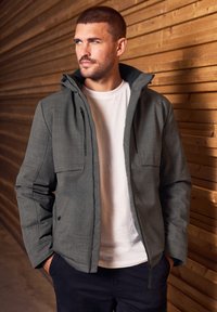 Mörkgrå, huvad jacka med en strukturerad yta, som har två bröstfickor och dragkedja framtill, bärs över en vit t-skjorta med rund halsringning.