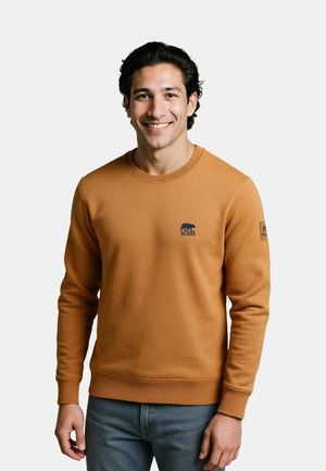 Jonge man die lacht, een bruine trui draagt met kleine logo's op borst en mouw, en blauwe spijkerbroek, staand tegen een effen lichte achtergrond.