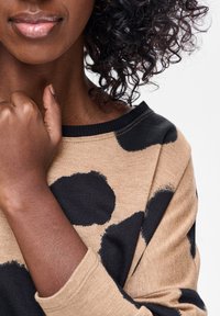 Brązowy sweter z dużymi czarnymi plamami, z szerokim dekoltem i czarnym prążkowanym wykończeniem. Widoczne kręcone włosy i skóra na zbliżeniu.