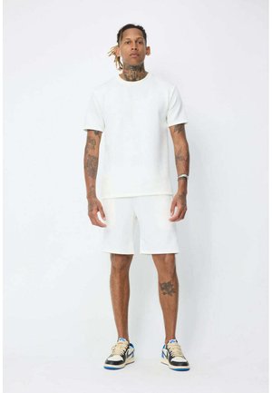 Homme tatoué portant un t-shirt blanc uni, un short blanc et des baskets noir et blanc avec des touches bleues, se tient devant un fond blanc.