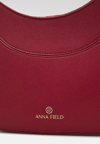 Punainen teksturoitu olkabag, jossa on kaareva yläosa ja kultainen "ANNA FIELD" -logo alaosassa sekä sileät saumat reunoilla.