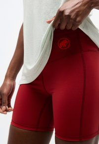 Κοντινό πλάνο ενός ατόμου που σηκώνει ένα ανοιχτό γκρι πουκάμισο για να αποκαλύψει κόκκινα ποδηλατικά σορτς με ψηλή μέση και λογότυπο μαμούθ στο ισχίο.