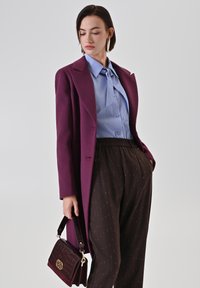Camicia button-up azzurro chiaro con cravatta, cappotto di lana borgogna, pantaloni scuri a fantasia e borsa testurizzata color bordeaux.