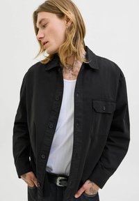 Person with long blond hair wearing an unbuttoned black shirt over a white tank top, black belt, and dark jeans, looking to the left.