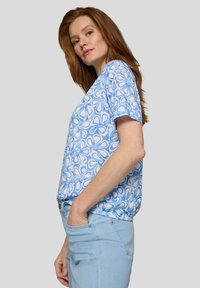 Bluse mit blauem Blumenmuster aus leichtem Stoff, mit kurzen Ärmeln und entspannter Passform, kombiniert mit hellblauen Jeans.