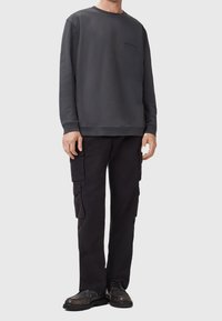 Homme portant un sweat-shirt gris foncé à manches longues et un pantalon cargo noir avec des chaussures noires, debout devant un fond uni gris clair.