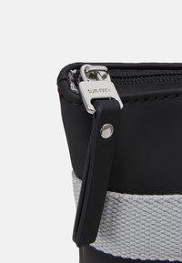 Pochette zippée noire avec quincaillerie en argent, dotée d'une sangle texturée grise. Le matériau semble lisse et flexible, avec des détails de couture visibles.