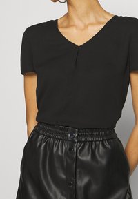 Top nero a maniche corte con scollo a V e una leggera piega frontale, abbinato a pantaloni neri in ecopelle a vita alta con una cintura arricciata.