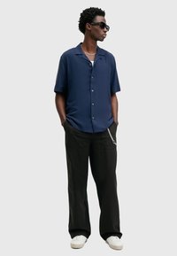 Marineblauwe korte mouwen button-up shirt met een relaxte pasvorm, gecombineerd met zwarte wijde broek en witte sneakers. Accessoires omvatten een zonnebril en een ketting.
