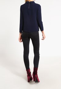 Blusa blu navy con collo e zip sulla schiena, abbinata a jeans skinny neri. La modella indossa stivaletti a blocco con tacco in velluto rosso.