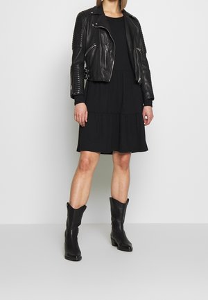 Chaqueta de moto de cuero negro con acentos acanalados, combinada con un vestido negro de capas y botas altas negras. Diseño y texturas minimalistas.