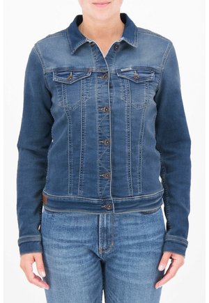 Femme portant une veste en jean bleu ajustée avec des fermetures à boutons et des poches à rabats sur la poitrine, par-dessus un jean bleu, debout sur un fond blanc.
