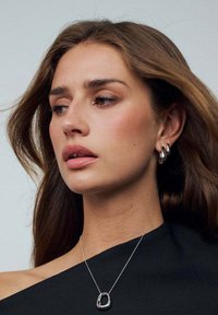 Modell med långt brunt hår som bär en svart off-shoulder topp, silverfärgade hoopörhängen och ett silverhalsband med en skulptural berlock.