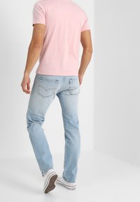 Camiseta ajustada de color rosa claro para hombres combinada con jeans de color azul claro. Los vaqueros están ligeramente desgastados. Las zapatillas deportivas tienen una base blanca con acentos negros.