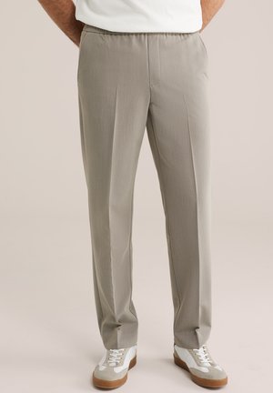 Világos bézs szabott nadrágok elülső hajtásokkal, fehér sportcipőkkel viselve, melyeken bézs részletek és gumi talpak találhatók.