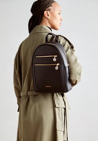 Mochila de cuero negro con acentos de cremallera dorada, dos bolsillos frontales, forma redondeada y un pequeño charm. Llevada en la espalda de una persona.