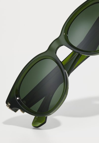 Zaļas pārmērīgi lielas saulesbrilles ar gludu apdari, aprīkotas ar apaļiem stikliem un biezām rāmī. Stikli ir tumši zaļi ar vieglu tonējumu.