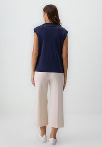 Donna in piedi di fronte a un muro bianco, indossa un top senza maniche blu navy, pantaloni a gamba larga beige chiaro e sneakers bianche.