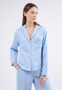 Světle modrý pyžamový set s růžovým lemováním. Tričko má kapsu s výšivkou mašle a knoflíky vpředu. Dlouhé rukávy.