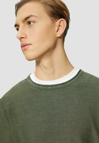 Sweatshirt vert olive avec un col rond, superposé sur une chemise à col rond blanche. Texture douce, coupe décontractée et détails de couture subtils.