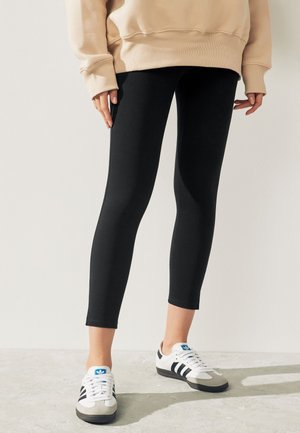 Person iført beige sweatshirt, sorte cropped leggings og hvide Adidas sneakers, stående på en lys gulv mod en ensfarvet væg.