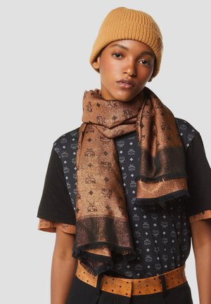 Jong persoon met een beige gebreide muts, metallic bruine sjaal met logo patroon, zwart-bruin logo T-shirt en bijpassende riem tegen een effen achtergrond.
