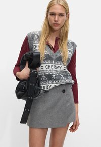 Sweter bez rękawów w szarym kolorze z białym wzorem roweru i napisem "CHERRY", zestawiony z ciemnoczerwonym topem z długim rękawem oraz szaro-skalowanym mini spódnicą. Trzyma czarną torbę.