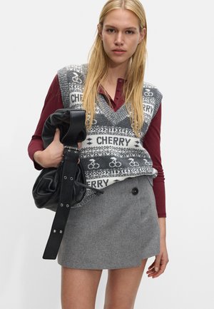Sweter bez rękawów w szarym kolorze z białym wzorem roweru i napisem "CHERRY", zestawiony z ciemnoczerwonym topem z długim rękawem oraz szaro-skalowanym mini spódnicą. Trzyma czarną torbę.
